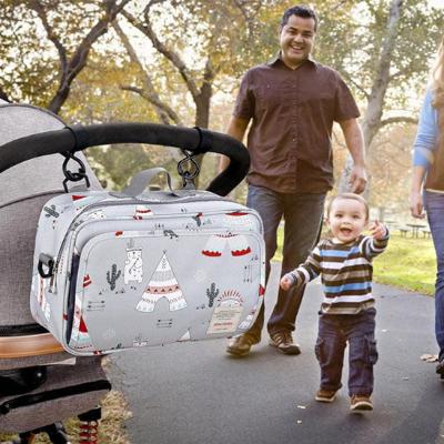 Sac à langer gris avec des motifs de tentes, cactus et bonhommes accroché à une poussette avec bébé et papa s'amusant librement. 